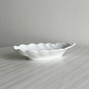 May include: A white ceramic serving dish shaped like a seashell with a scalloped edge and a textured, embossed pattern on one end. The dish has a shallow, oval-shaped basin and a smooth, glossy finish, presenting a clean and simple aesthetic.
