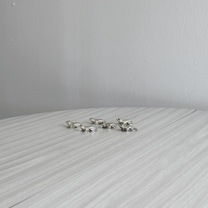 May include: Several small, silver frog figurines are displayed on a light grey, wood-effect surface. The frogs are in different poses, with detailed features. The background is a plain, light grey wall.