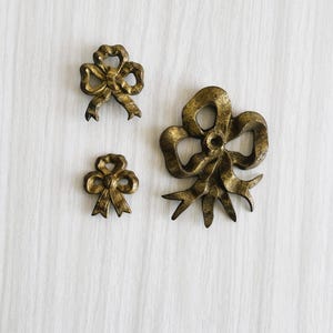 Könnte beinhalten: Drei antike, goldfarbene Metall-Schleifenornamente in verschiedenen Größen. Die Schleifen weisen aufwendige Details und eine abgenutzte Oberfläche auf, was auf ein gealtertes Aussehen hindeutet. Sie sind auf einer weißen, strukturierten Oberfläche angeordnet, wahrscheinlich zu Dekorationszwecken.