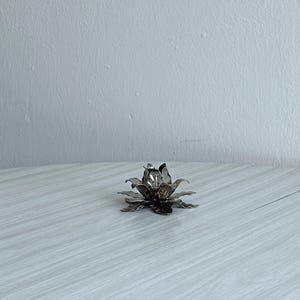 Könnte beinhalten: Ein silberfarbener Kerzenhalter aus Metall in Form einer blühenden Lotusblume. Das Objekt besteht aus mehreren Schichten spitzer, texturierter Blütenblätter, die von einem Mittelpunkt ausstrahlen und eine symmetrische, mehrstufige florale Silhouette bilden.
