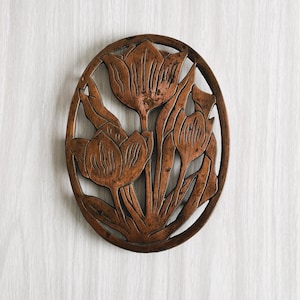 Puede incluir: Un salvamanteles de metal de color cobre con un diseño recortado de tres tulipes. El salvamanteles es redondo y tiene un acabado liso y pulido.