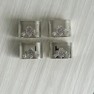 Peut inclure: Un ensemble de quatre ronds de serviette en métal argenté, chacun présentant une finition polie et un motif en relief représentant une grappe de raisin avec des feuilles au centre. Les anneaux ont une forme cylindrique lisse.