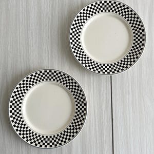Op de afbeelding: Twee ronde keramische borden met een effen gebroken wit midden en een brede rand versierd met een herhalend zwart-wit geblokt patroon. De randen van de borden zijn afgewerkt met een dunne, neutraal gekleurde rand voor een strak ontwerp.