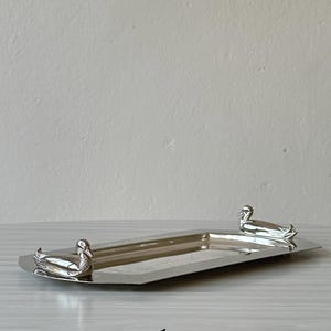 Peut inclure: Un plateau rectangulaire en métal argenté doté de deux figurines de canards sculptées fixées aux extrémités opposées du rebord. Le plateau présente une surface lisse et réfléchissante ainsi qu'un bord surélevé, avec le chiffre 1 inscrit en dessous.