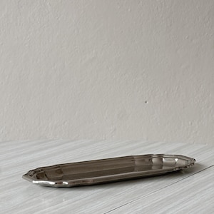 Peut inclure: Un plateau rectangulaire long et étroit en métal argenté, doté de coins arrondis et d'un bord surélevé festonné. Le plateau présente une surface lisse et réfléchissante avec une finition polie et un centre légèrement creusé, présenté horizontalement.