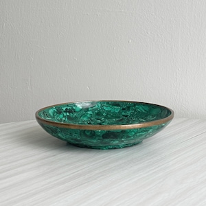 Può includere: Una ciotola decorativa con un vivace motivo verde malachite e un bordo color oro. La ciotola ha una forma poco profonda e arrotondata e poggia su una superficie chiara. Questo è un oggetto decorativo.