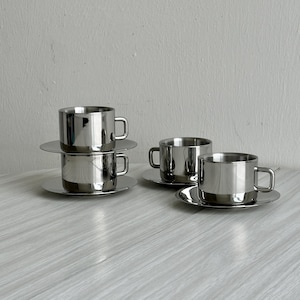 Puede incluir: Un juego de cuatro tazas de café y platillos de acero inoxidable. Dos tazas están apiladas y las otras están dispuestas sobre la mesa. Las tazas tienen un diseño simple y moderno con un asa. Los platillos son redondos y planos.