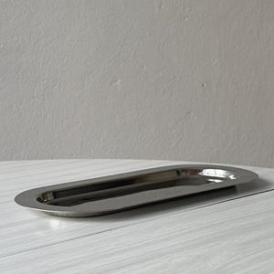 Pode incluir: Uma bandeja de metal prateado polido com formato oval alongado e superfície lisa e reflexiva. A bandeja apresenta uma borda larga e plana que se curva suavemente em direção ao centro raso e rebaixado, criando um perfil elegante.