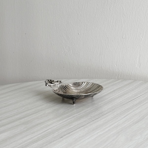 May include: A small, silver-toned dish with a shell-like design. The dish has a scalloped edge and rests on three small feet. The interior features a textured, ridged pattern. A decorative element adorns one side of the dish.