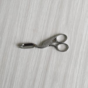 May include: A silver-colored, decorative sewing scissor with a bird-like design. The scissor features a blade on one end and two finger holes on the other. The scissor is laying on a white, textured surface.