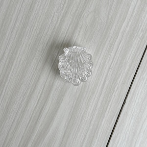 May include: A clear, seashell-shaped decorative object. The object is made of a transparent material and features detailed ridges, resembling a real seashell. The object is placed on a light gray surface.