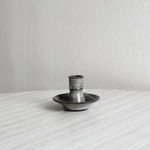 May include: A ceramic candlestick holder with a grey and brown glaze. The candlestick holder has a small, round base and a cylindrical centre for holding a candle. The piece is on a white surface.