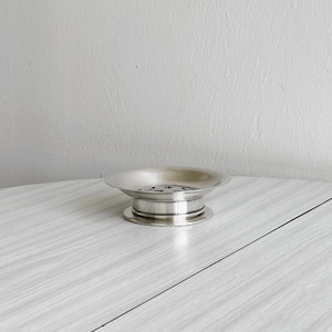 Puede incluir: Un jabonera plateada con un cuenco circular y poco profundo y una base elevada y escalonada. La jabonera se encuentra sobre una superficie clara con vetas de madera contra una pared blanca lisa. La jabonera es probablemente de metal.