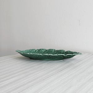 Op de afbeelding: Een groene, bladvormige serveerschaal van keramiek. De schaal heeft een oppervlak met textuur, gedetailleerde nerven en een geschulpte rand. De kleur is een diep, rijk groen en de schaal staat tegen een neutrale achtergrond.