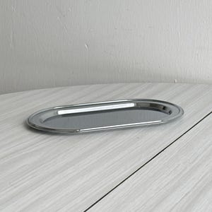 Könnte beinhalten: Ein ovaler Servierteller aus poliertem Edelstahl. Das Tablett hat eine reflektierende Oberfläche und einen leicht erhöhten Rand. Das Tablett ist ca. 30 cm lang und 15 cm breit.