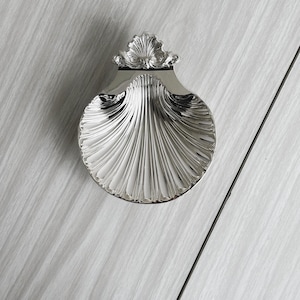 Könnte beinhalten: Eine silberfarbene, muschelförmige Dekoschale. Die Schale hat ein detailliertes, geriffeltes Design und eine dekorative Oberseite. Die Schale besteht aus einem glänzenden, reflektierenden Material und ist vor weißem Hintergrund platziert.