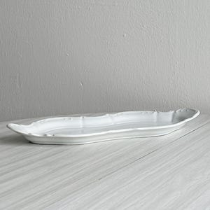 Peut inclure: Un long plat de service en céramique blanche en forme de feuille. Le plat a un bord festonné et repose sur une surface en bois de couleur claire. L'arrière-plan est un mur uni gris clair.