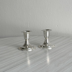 May include: A pair of polished silver-toned metal candle holders featuring a flared base and a matching flared top rim. Each holder has a smooth, reflective surface and a decorative beaded edge detail around the circumference of the base and top.