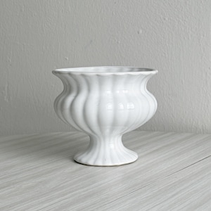 Pode incluir: Uma taça de pedestal em cerâmica branca com acabamento brilhante e um padrão de nervuras verticais distinto. O recipiente tem uma borda larga e alargada que se curva para fora, estreitando-se para um pescoço antes de uma base circular.