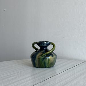 Può includere: Un piccolo vaso in ceramica verde e blu con due manici. Il vaso ha una smaltatura screziata e un corpo arrotondato.