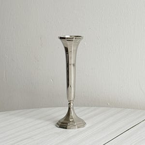 May include: A silver-coloured bud vase with a flared top and a faceted design. The vase has a slender, elongated body and a pedestal base. The vase is made of metal and is displayed on a white surface.