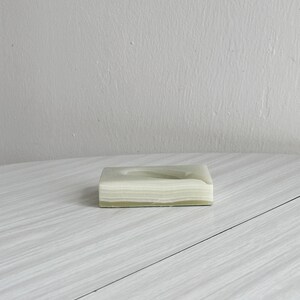 May include: A rectangular, light green soap dish with a layered, striped design. The soap dish is made of a solid material and sits on a light gray surface.