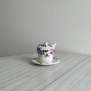 May include: A small white ceramic sugar bowl with a lid and a matching saucer. The bowl and saucer are decorated with a floral pattern of purple flowers and green leaves.