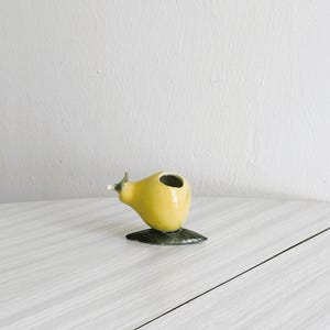 Puede incluir: Un pequeño jarrón de cerámica amarilla con forma de pera, con una base de hoja verde oscuro y una pequeña abertura en la parte superior. El jarrón tiene un acento blanco en la parte superior y está sobre un fondo blanco.