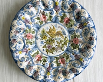 May include: A decorative ceramic plate with a floral and bird design. The plate features a blue rim and a central image of a yellow bird surrounded by flowers. The plate's design includes pink, blue, and green floral patterns.