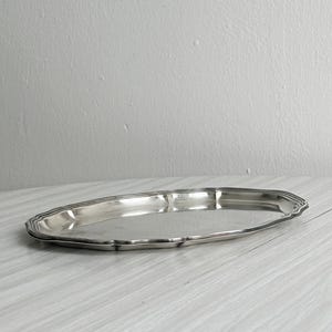 Puede incluir: Una bandeja de servicio ovalada de tono plateado con una superficie pulida y reflectante y un borde decorativo festoneado. La bandeja presenta un centro liso y plano y un borde elevado de varias capas que sigue la forma curva y alargada.