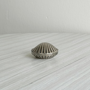 Könnte beinhalten: Kleine, silberfarbene Schmuckschatulle in Form einer Muschel. Die Schatulle hat ein geripptes Design auf der Ober- und Unterseite mit einem detaillierten Rand. Die Schatulle ist geschlossen und liegt auf einer hellen Oberfläche.