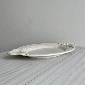 Può includere: Un piatto da portata ovale bianco con dettagli ornati e in rilievo sui manici. Il piatto ha una superficie leggermente strutturata ed è appoggiato su un tavolo di legno chiaro. Lo sfondo è un muro grigio chiaro e uniforme.