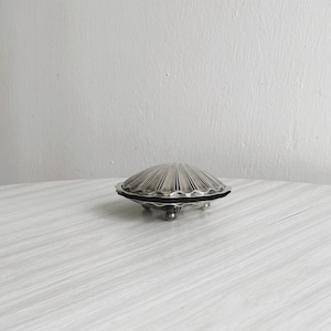 May include: A silver-colored, shell-shaped trinket dish with a ridged design. The dish sits on three small, ball-shaped feet and has a black rim. The lid is closed, and the dish is on a light-colored, wood-grain surface.