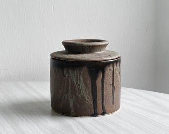 Può includere: Una burriera in ceramica marrone con coperchio. Il contenitore cilindrico ha una glassa marrone scuro con gocciolature e il coperchio è della stessa tonalità. Il piatto è progettato per conservare il burro e ha un'estetica rustica e artigianale.
