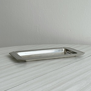 May include: A rectangular silver-toned metal tray featuring a smooth, reflective surface and a wide, flat rim with beveled edges. The tray has a shallow basin and a clean, polished finish, presenting a simple and functional design for holding small items.