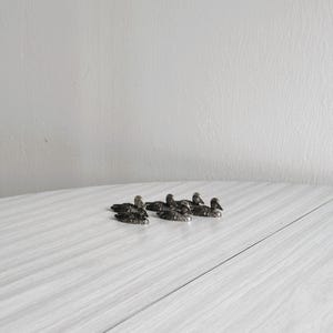 May include: A set of six silver duck place card holders. The ducks are arranged in a line on a white wooden surface.