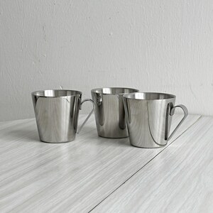 Puede incluir: Tres tazas de acero inoxidable con asas anguladas están dispuestas sobre una superficie de madera de color claro. Las tazas tienen un diseño simple y moderno con un acabado pulido y reflectante. El fondo es una pared lisa de color blanquecino.