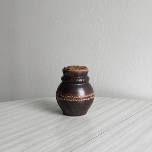Può includere: Un vaso in ceramica marrone scuro con un coperchio intrecciato. Il vaso ha una fascia decorativa intorno al centro.