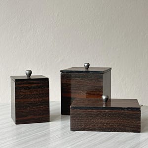May include: Three dark brown wooden boxes with lids, each featuring a small, rounded silver-toned metal knob handle. The set includes one tall rectangular box, one square box, and one low, wide rectangular box, all with a visible wood grain finish.