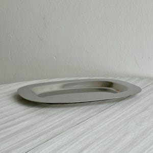 Puede incluir: Una bandeja ovalada de acero inoxidable. La bandeja tiene una superficie pulida y reflectante y un borde ligeramente elevado. La bandeja está vacía y se encuentra sobre una superficie de madera gris claro. El fondo es una pared gris claro.