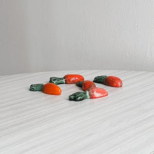 Può includere: Una collezione di statuette a forma di carota in ceramica. Ogni statuetta presenta un corpo di carota arancione vibrante e una parte superiore verde fogliata. Le statuette sono disposte su una superficie di colore chiaro.