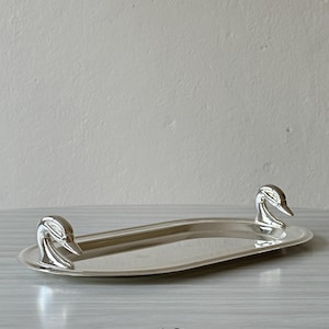 May include: An oval silver-toned metal tray featuring two sculpted duck head handles on opposite ends. The tray has a smooth, reflective surface and a raised rim. The number 10 is printed in black below the tray on the surface.