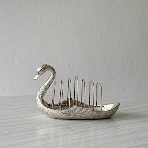 May include: A silver-toned metal toast rack shaped like a swan. The piece features a textured, feathered body design and a series of vertical metal dividers extending from the back to hold slices of toast in an upright position.
