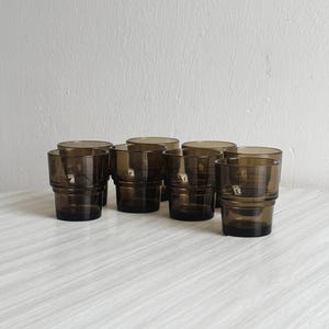 May include: A collection of eight short, stackable drinking glasses. The glasses are made of a dark, smoky-brown glass. Each glass has a thick, cylindrical shape with a ridged design around the middle. The glasses are arranged on a light-colored surface.