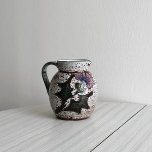 Puede incluir: Una jarra de cerámica blanca con un diseño floral marrón, verde y rosa. La jarra tiene un esmalte moteado y un cuerpo redondeado.