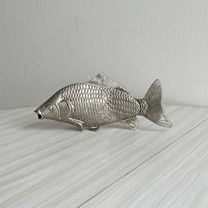 May include: A silver fish-shaped bottle opener. The opener has detailed scales, fins, and a tail. The fish is on a light-coloured wooden surface.