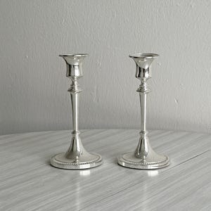 Peut inclure: Une paire de bougeoirs en métal argenté poli avec une tige fine, une bobèche évasée au sommet et une base circulaire ornée d'un bord perlé. Ces supports de bougies métalliques présentent une finition lisse et réfléchissante.
