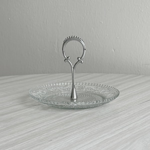 Peut inclure: Un plateau de service en verre transparent avec un motif texturé et gaufré sur le bord et au centre. Le plateau est doté d'une poignée centrale en métal argenté avec un design en boucle perlée sur le dessus pour faciliter le transport.