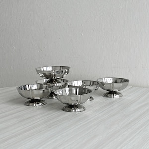 May include: A set of six silver-colored dessert bowls. Each bowl has a scalloped rim and sits on a pedestal base. The bowls are arranged on a light-colored surface against a neutral background.