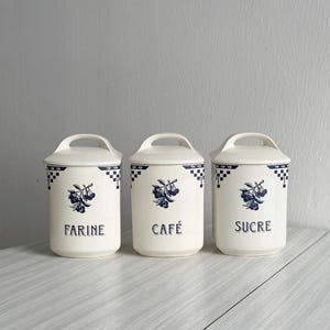 Puede incluir: Tres botes de cerámica blanca con diseños azules y blancos. Los botes están etiquetados con "Farine", "Cafe" y "Sucre".